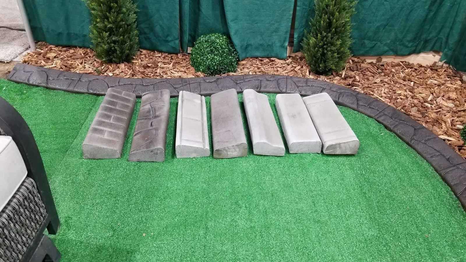 Seven concrete curb samples are displayed on artificial turf beside a curved stamped demonstration edge.