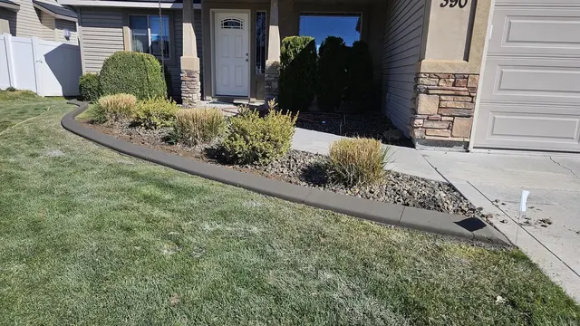 A dark smooth curb frames shrubs, ornamental grasses and stone at a house entrance.