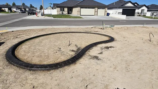 A dark stamped concrete edge encloses a rounded planting island in a newly graded yard.