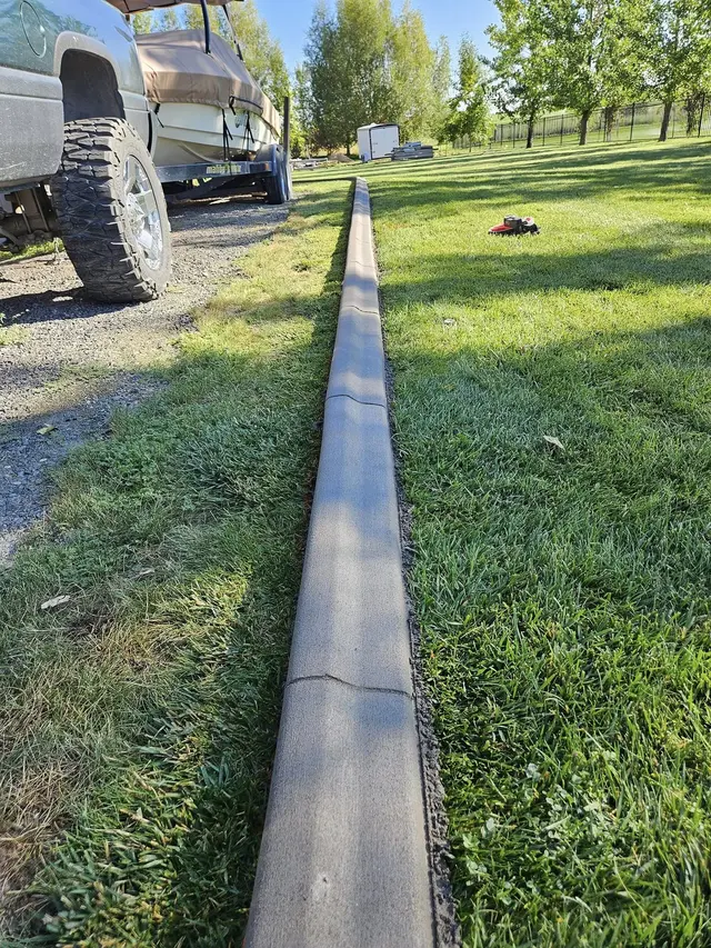 A portrait view looks along a straight gray border between a lawn and a gravel parking strip.