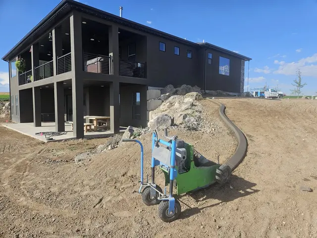 A green curbing machine sits beside a new concrete edge on a graded slope near a dark house.