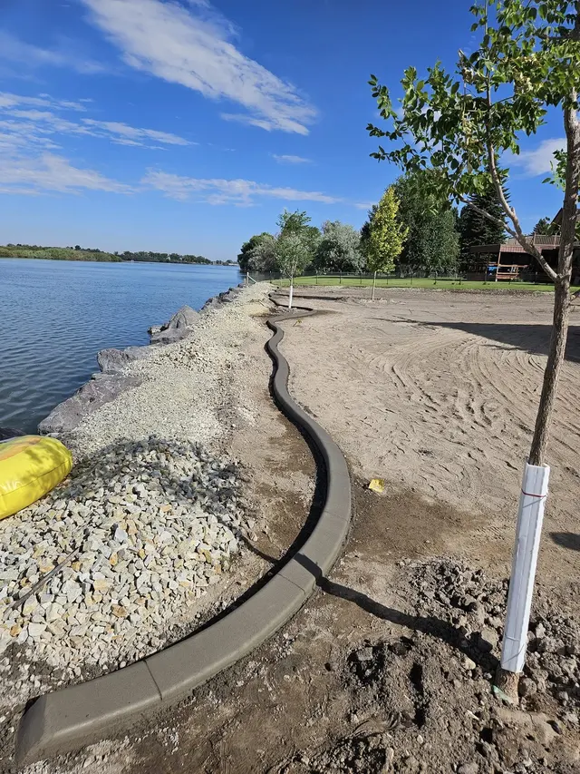A portrait view follows a gray curb between a gravel lakeshore and bare garden soil.