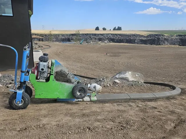 A green curbing machine rests beside a newly formed curved edge and large rocks.
