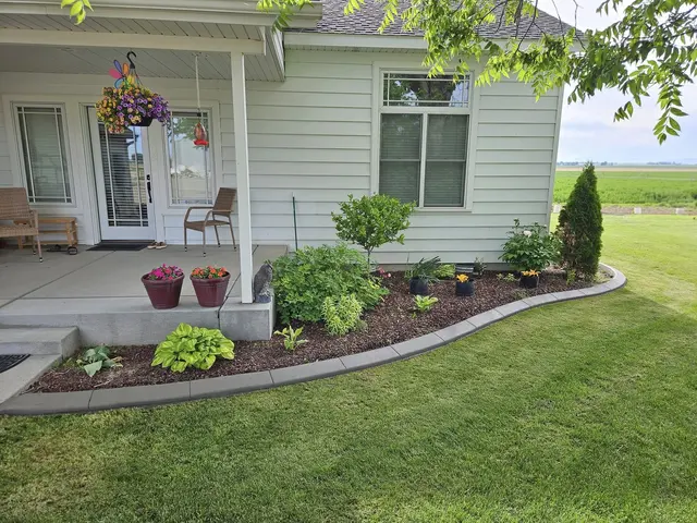 A smooth gray edge encloses shrubs, flowers, and several nursery pots beside a front porch.