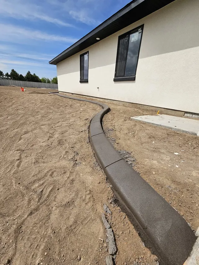 A gray concrete edge sweeps alongside a white house through freshly graded soil.