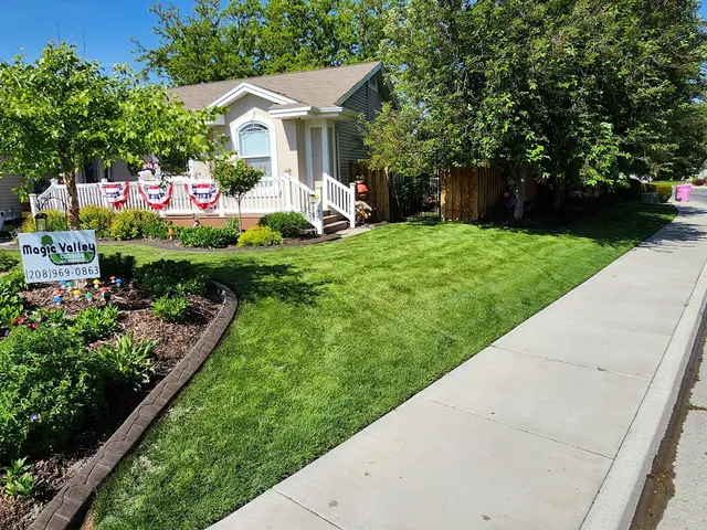 A curving garden border frames a lush lawn and flower beds beside a house with porch bunting.