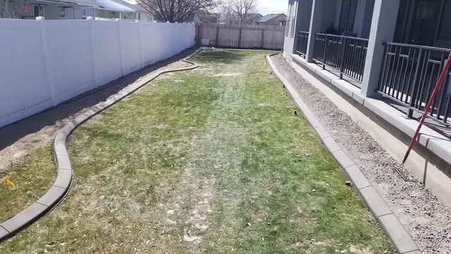A wide view shows gray curbing running along both sides of a narrow lawn beside a porch and fence.