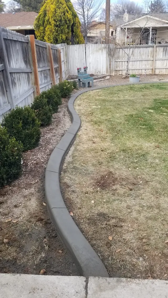 A flowing gray border separates lawn and a narrow shrub bed beside a wooden fence.