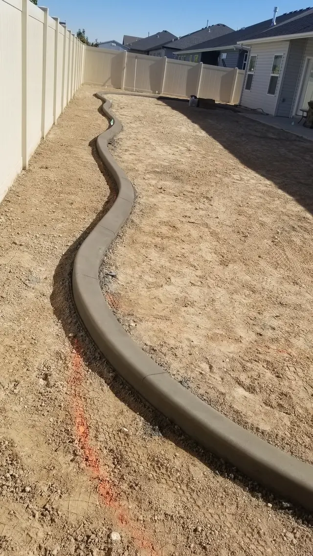 A dark smooth edge follows a long wavy line through bare soil beside a light fence.