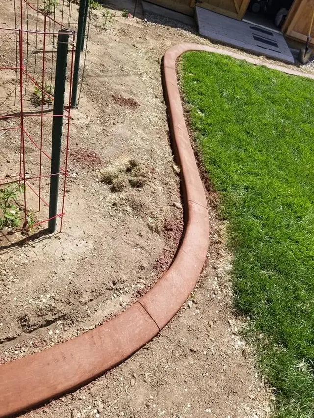 A close portrait follows a warm red-brown concrete edge between green grass and bare soil.