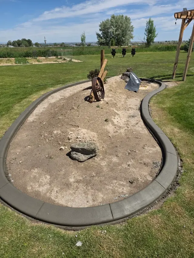Long organic island bounded by fresh dark concrete in a green yard, with bare soil and rustic ornaments inside.
