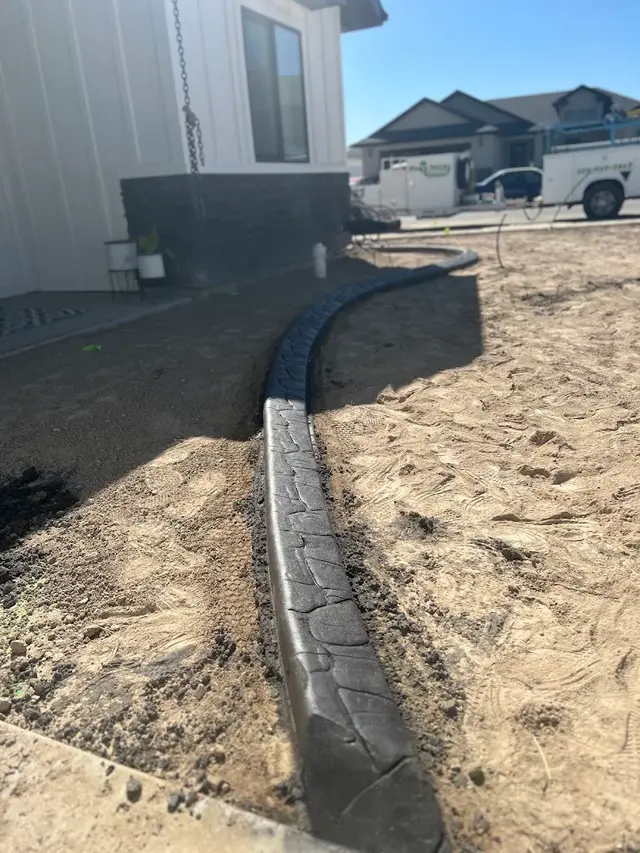 Dark stamped concrete curb follows the side of a new house through exposed soil.