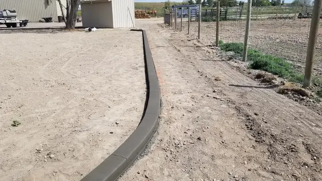Long straight-to-curved gray boundary beside a fenced rural growing area and prepared soil.