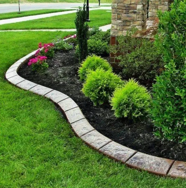 Pale stamped sweeping curb around a dark mulched bed, bright shrubs, flowers, and a stone column.