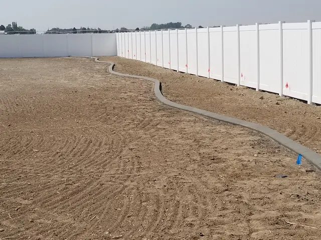 Long fresh gray curve across a graded bare-soil backyard along a white fence.
