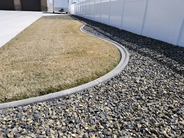 Long pale S-curve between dormant lawn and a dark rock bed along a white fence.