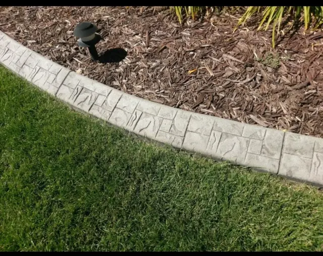 Close view of a pale stamped curb separating green lawn and brown mulch.