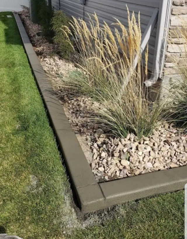 Straight gray curb with a crisp right-angle turn around grasses in a rock bed beside a house.